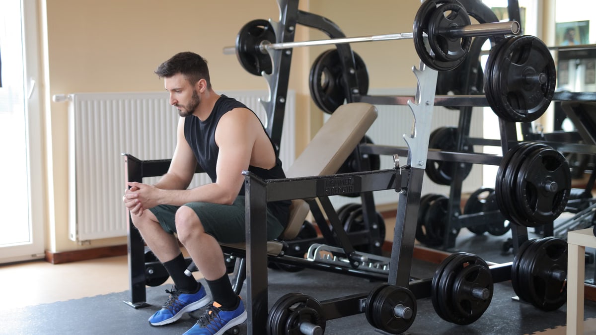 A man takes a break at the gym sitting beside a fully loaded barbell, showcasing strength.